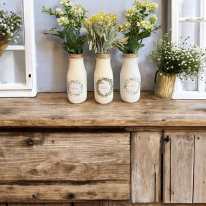 May include: Three white milk glass bottles with floral wreaths and the words "willow", "home", and "welcome" on them. The bottles are filled with different types of flowers and are sitting on a wooden surface.
