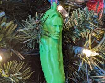 Puede incluir: Un adorno navideño verde con forma de pepinillo con una cara humana. El adorno está colgado de un árbol de Navidad.