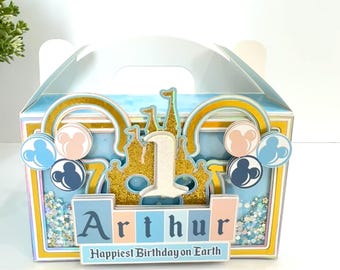 May include: A blue and gold party favor box with a castle, Mickey Mouse ears, and the number one. The box has glitter accents and says "Arthur Happiest Birthday on Earth".