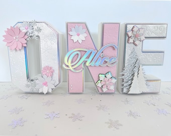May include: White and pink cardboard letters spelling "ONE" with silver glitter and snowflake embellishments. The letter "N" has the name "Alice" written in iridescent letters.