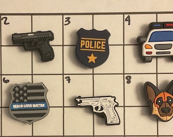 May include: Eight different shoe charms, including a pair of sunglasses, two guns, a police badge, a police car, a badge with the words "Black Lives Matter", a German Shepherd dog, and a thin blue line flag.