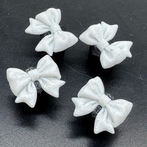 May include: Four white bow-shaped accessories. Each bow is detailed with textured patterns and sits atop a dark, cylindrical base. The bows are arranged against a dark background.