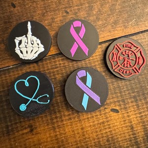 May include: A collection of five black circular objects with various designs. One features a white skeleton hand giving the middle finger, another a pink ribbon, and a third a red firefighter emblem. The remaining two have a blue stethoscope and a purple and blue ribbon.
