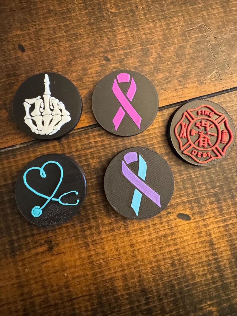 May include: A collection of five black circular objects with various designs. One features a white skeleton hand giving the middle finger, another a pink ribbon, and a third a red firefighter emblem. The remaining two have a blue stethoscope and a purple and blue ribbon.