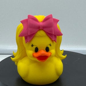 Può includere: Un'anatra di gomma gialla con capelli biondi e un fiocco rosa. L'anatra ha occhi neri e un becco arancione. L'anatra è su una superficie nera.
