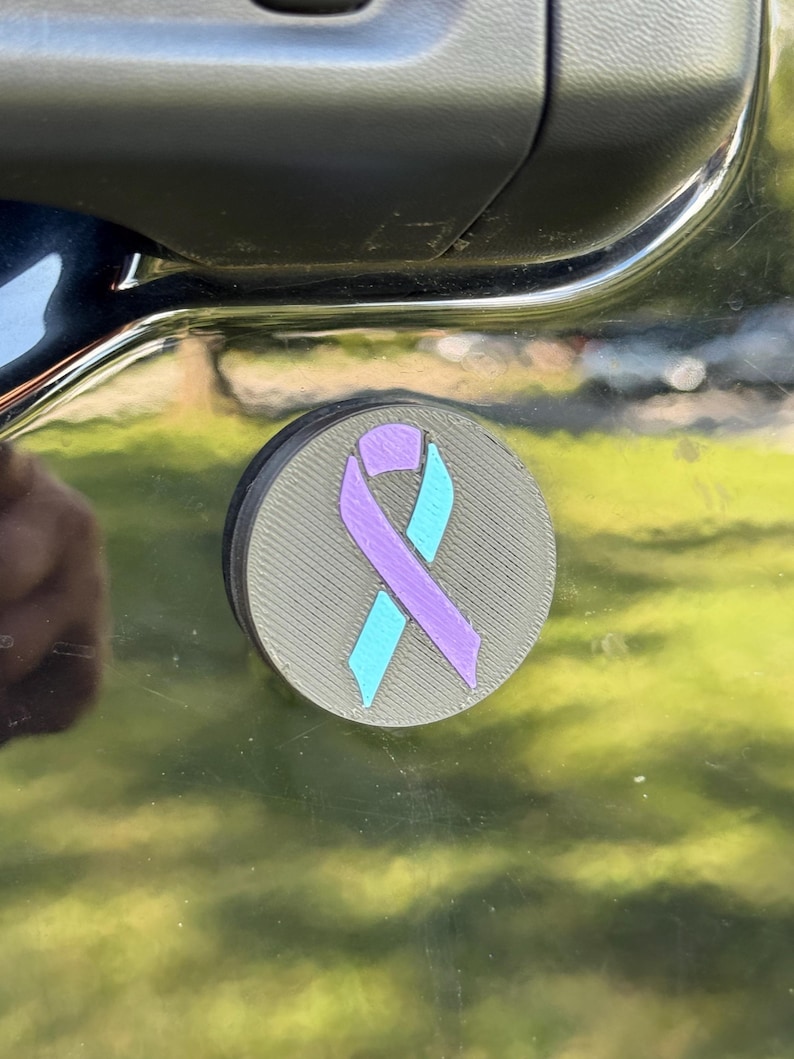 May include: A round, gray car magnet featuring a purple and teal awareness ribbon. The magnet is affixed to a dark-colored vehicle door, reflecting a blurred green and yellow background.