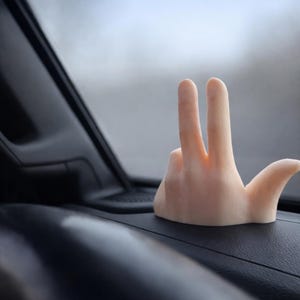 May include: A light-colored, hand-shaped car accessory with two fingers raised in a peace sign. The accessory is placed on a black dashboard, with a blurred background of a car interior and exterior.