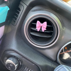 May include: A pink fabric bow car air vent clip. The bow is centered in the vent, adding a decorative touch to the car's interior. The dashboard is black with silver accents.