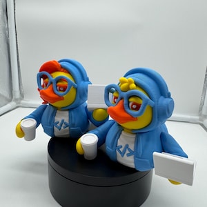 May include: Two yellow duck figurines wearing blue hoodies, glasses, and headphones. Each duck holds a white cup and a white rectangle. The hoodies have a white shirt with a code symbol. The ducks are on a black platform.