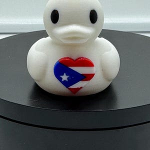 May include: A white rubber duck with a red, white, and blue heart-shaped design on its chest. The heart features the Puerto Rican flag.
