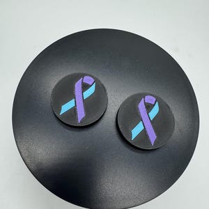 May include: Two round, black patches with a textured surface. Each patch features a light blue and purple ribbon design. The patches are displayed on a black, circular surface.