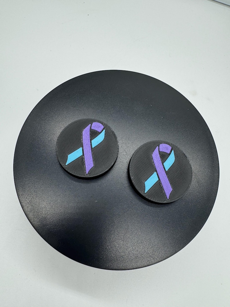 May include: Two round, black patches with a textured surface. Each patch features a light blue and purple ribbon design. The patches are displayed on a black, circular surface.