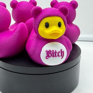 May include: A group of magenta rubber duck toys, one wearing a bear costume with the word "Bitch" on its chest. The duck's beak is yellow, and the costume has a hood with bear ears. The ducks are on a black surface.