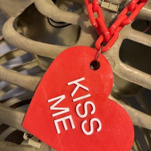 May include: A red heart-shaped tag with the words "Kiss Me" in white lettering, attached to a red plastic chain.