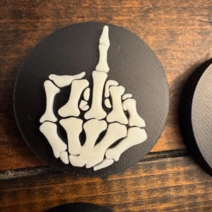 May include: A black, round coaster featuring a white, skeletal hand with the middle finger raised. The coaster is made of a dark material and is set against a wooden surface. The design is a bold, graphic statement.