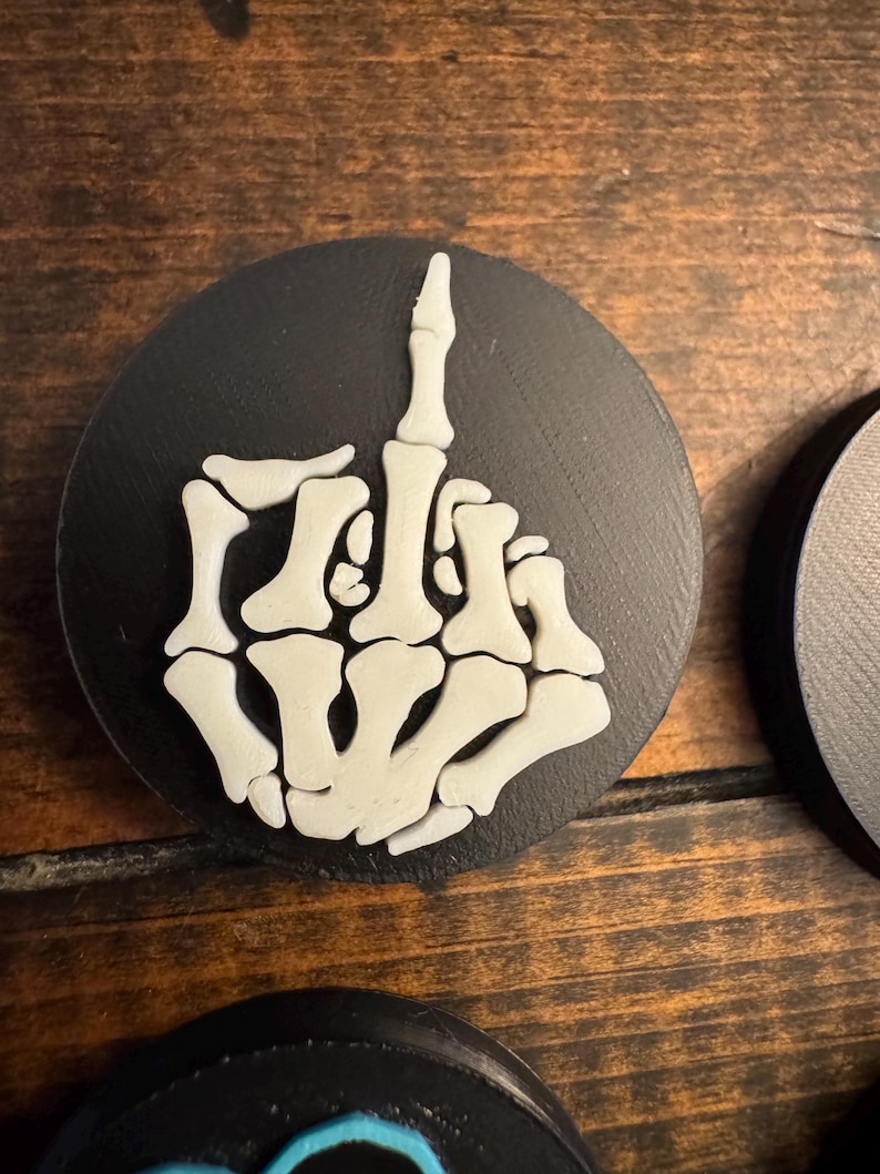May include: A black, round coaster featuring a white, skeletal hand with the middle finger raised. The coaster is made of a dark material and is set against a wooden surface. The design is a bold, graphic statement.