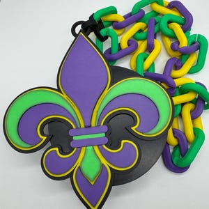 May include: A purple, green, and yellow fleur-de-lis emblem with a black backing and a matching chain. The emblem is a symbol of the Mardi Gras celebration. The chain is made of interlocking links in green, yellow, and purple.
