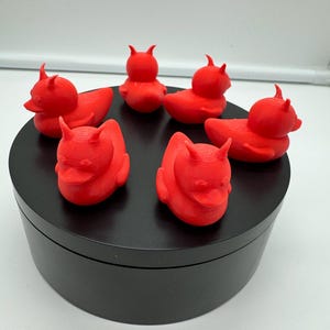 May include: Six red rubber duckies with devil horns, arranged in a circle on a black surface.