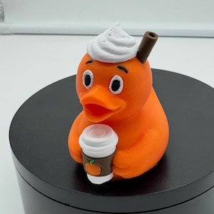 May include: Orange duck figurine with a white whipped cream topping and a brown cinnamon stick. The duck is holding a small coffee cup with a pumpkin graphic. The figurine is on a black surface.