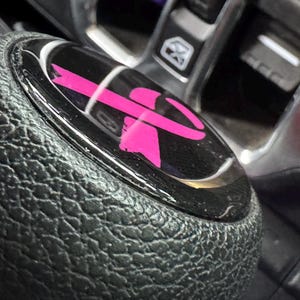 May include: A black textured gear shift knob featuring a circular glossy black insert on top. The insert displays a bright pink ribbon graphic centered on the surface. The knob is positioned within a vehicle interior near a console panel.