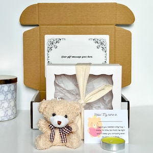 May include: A gift box with a teddy bear, a card, and a candle. The teddy bear is light brown with a checkered bow. The card reads "Hello! My name is..." and "Your gift message goes here."