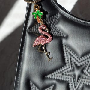 May include: A pink rhinestone flamingo charm with gold-colored accents and a green and brown palm tree charm, attached to a gold keyring. The charm hangs from a black handbag with star-shaped embossed details.