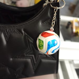 May include: A black handbag with a quilted star pattern features a spherical keychain attached to its silver metal ring. The keychain is a white ball decorated with red, blue, and green graphic panels, suspended by a short silver chain link.