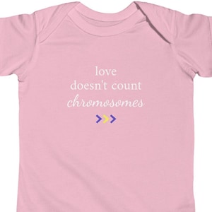 Peut inclure: Un body pour bébé rose clair avec le texte blanc "love doesn't count chromosomes" imprimé sur la poitrine. Sous le texte se trouvent trois petits symboles en forme de chevron de couleur jaune et violette, alignés horizontalement.
