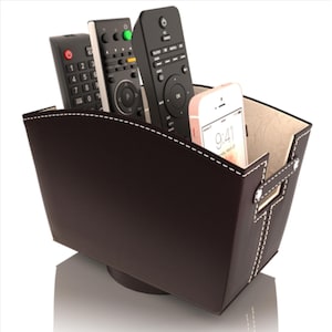 May include: A dark brown leatherette storage bin with white stitching, holding several black remote controls and a white and pink smartphone. The bin has a rectangular shape with handles and a light-colored interior.