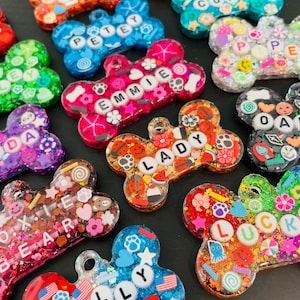 May include: A collection of colorful, bone-shaped pet tags with various names and decorative elements. The tags are made of resin and glitter, with names like "PETEY", "EMMIE", and "LADY" spelled out in white letters. The tags feature various designs, including flowers, stars, and paw prints.