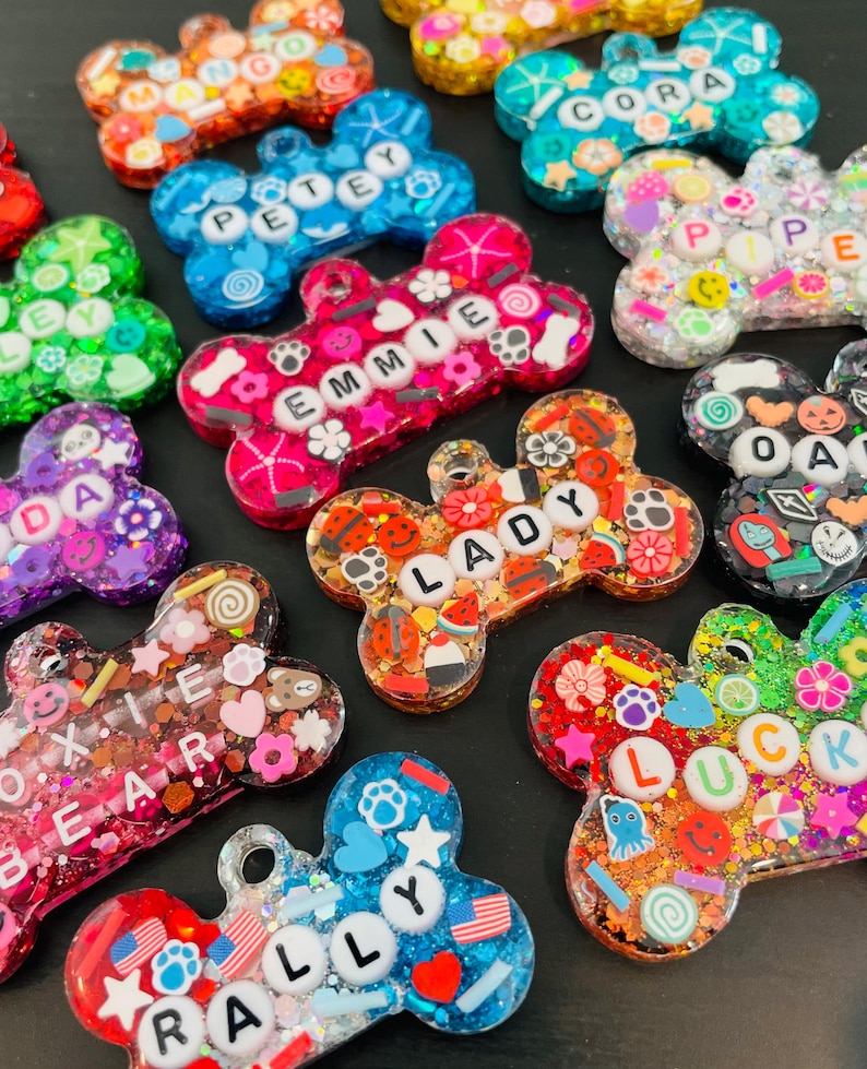 May include: A collection of colorful, bone-shaped pet tags with various names and decorative elements. The tags are made of resin and glitter, with names like "PETEY", "EMMIE", and "LADY" spelled out in white letters. The tags feature various designs, including flowers, stars, and paw prints.