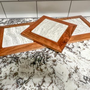 May include: Three square wooden coasters with a light gray marble inlay. The coasters are arranged in a stack, with the largest on the bottom and the smallest on top. The coasters are on a white and gray speckled countertop.