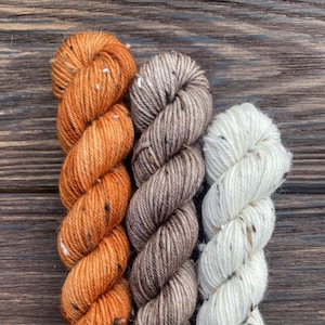Könnte beinhalten: Drei Garnknäuel in Orange-, Braun- und Weißtönen, jeweils mit gesprenkelten Details. Das Garn ist vertikal vor einem dunklen Holzmaserungshintergrund angeordnet und zeigt die Textur- und Farbvariationen.