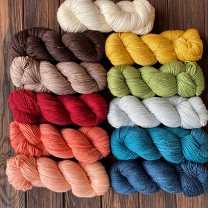 May include: A selection of colourful yarn skeins in shades like cream, brown, yellow, green, red, white, peach, teal, and navy blue. The yarn is displayed on a dark wooden surface, highlighting the different colours and textures.