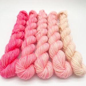 Könnte beinhalten: Eine Sammlung von Mini-Garnknäueln in Rosa- und Cremetönen. Das Garn ist in einem Farbverlauf angeordnet, der von einem leuchtenden Pink zu einem weichen, hellen Creme übergeht. Die Knäuel sind fest gewickelt und wirken weich.