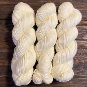 May include: Three skeins of white yarn, arranged in a row on a wooden surface.