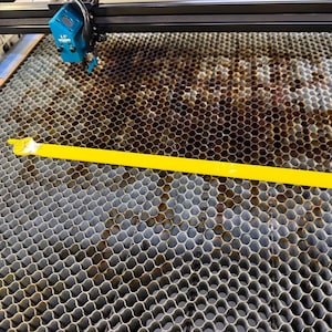 Puede incluir: Una regla de plástico amarilla descansa sobre una superficie metálica de panal, parte de una máquina de corte por láser. La máquina tiene un cabezal láser azul y un marco negro. La superficie de panal está manchada con residuos oscuros.