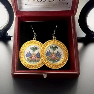 May include: A pair of gold earrings with a circular design featuring the coat of arms of Haiti. The earrings are in a red velvet box.