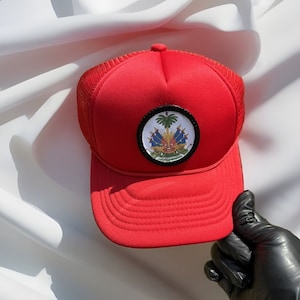 Op de afbeelding: Rode truckercap met een zwart-witte geborduurde patch van het Haïtiaanse wapenschild. De patch toont een palmboom, een kanon en een banner met de woorden "L'Union Fait La Force".