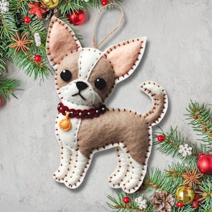 Może przedstawiać: Choinkowa ozdoba z filcu w kształcie chihuahua. Ozdoba jest brązowa, biała i czerwona z złotym dzwonkiem wok&oacute;ł szyi. Ozdoba jest otoczona zieloną gałązką sosny z czerwonymi i złotymi ozdobami.