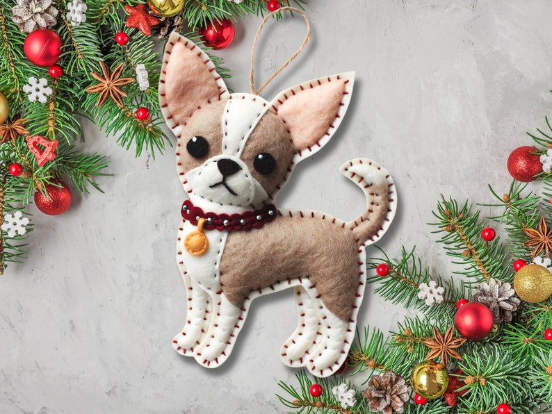 Może przedstawiać: Choinkowa ozdoba z filcu w kształcie chihuahua. Ozdoba jest brązowa, biała i czerwona z złotym dzwonkiem wok&oacute;ł szyi. Ozdoba jest otoczona zieloną gałązką sosny z czerwonymi i złotymi ozdobami.