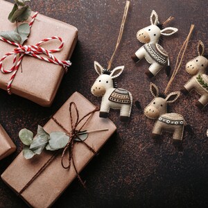 May include: A collection of six felt donkey ornaments with different patterns and colors. The ornaments are hanging from twine and are displayed on a brown background with wrapped gifts.