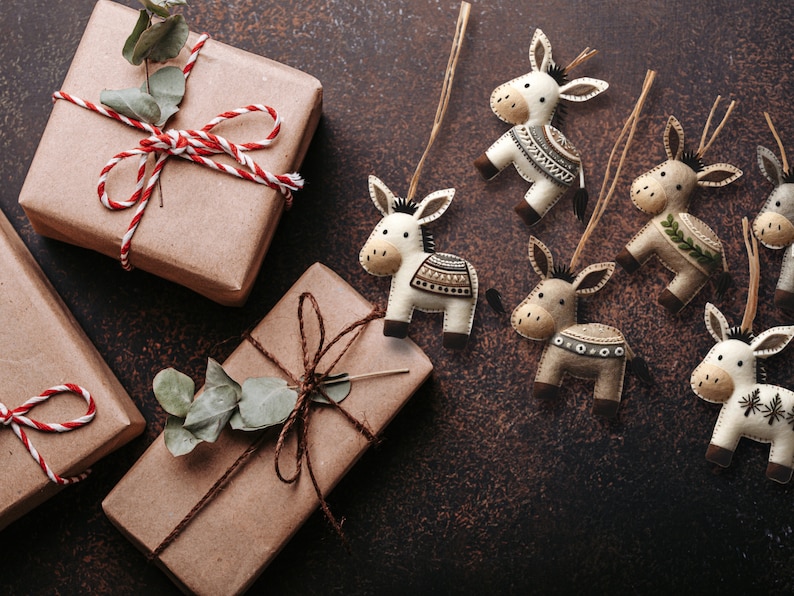 May include: A collection of six felt donkey ornaments with different patterns and colors. The ornaments are hanging from twine and are displayed on a brown background with wrapped gifts.