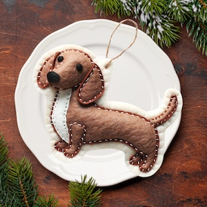 Może przedstawiać: Ozdoba w kształcie szczeniaka rasy dachshund z filcu w kolorze brązowym, z białymi szwami i białym filcowym paskiem na klatce piersiowej. Ozdoba jest zawieszona na białym sznurku i leży na białym talerzu.