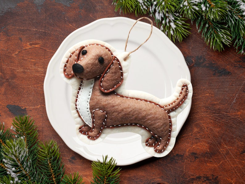 Może przedstawiać: Ozdoba w kształcie szczeniaka rasy dachshund z filcu w kolorze brązowym, z białymi szwami i białym filcowym paskiem na klatce piersiowej. Ozdoba jest zawieszona na białym sznurku i leży na białym talerzu.