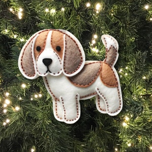 Może przedstawiać: Choinkowa ozdoba z filcu w kształcie beagle'a. Pies jest biały z brązowymi i beżowymi plamami. Ma brązowy nos i czarne oczy.