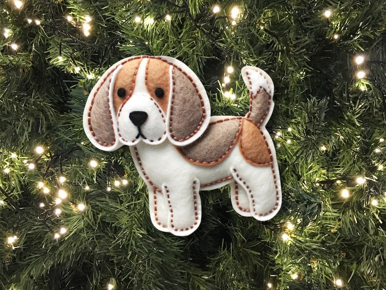 Może przedstawiać: Choinkowa ozdoba z filcu w kształcie beagle'a. Pies jest biały z brązowymi i beżowymi plamami. Ma brązowy nos i czarne oczy.