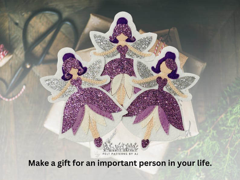 Pu&ograve; includere: Tre ornamenti di fata in feltro viola con ali di glitter argentate e dettagli bianchi. Le fate indossano abiti viola e hanno i capelli viola. Il testo "FELT PATTERNS BY AJ" &egrave; nella parte inferiore dell'immagine.