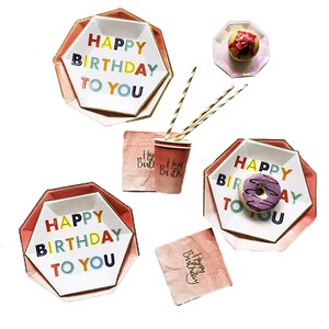 Könnte beinhalten: Ein Set aus rosa und weißen Papptellern mit goldener Verzierung und dem Text "Happy Birthday To You" in bunten Buchstaben. Die Teller sind mit einem Donut und einem Cupcake verziert. Außerdem gibt es rosa Servietten mit dem Text "Happy Birthday" und eine rosa Tasse mit dem Text "Happy Birthday".