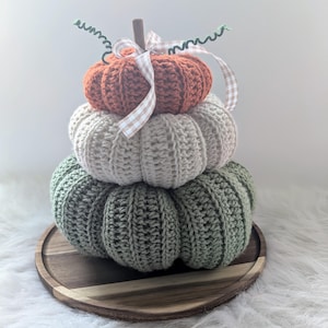 May include: A stack of three crocheted pumpkins in shades of orange, cream, and green, arranged on a wooden tray. The top pumpkin has a brown stem and is adorned with a gingham ribbon and green curly accents. The pumpkins are a fall-themed decorative item.